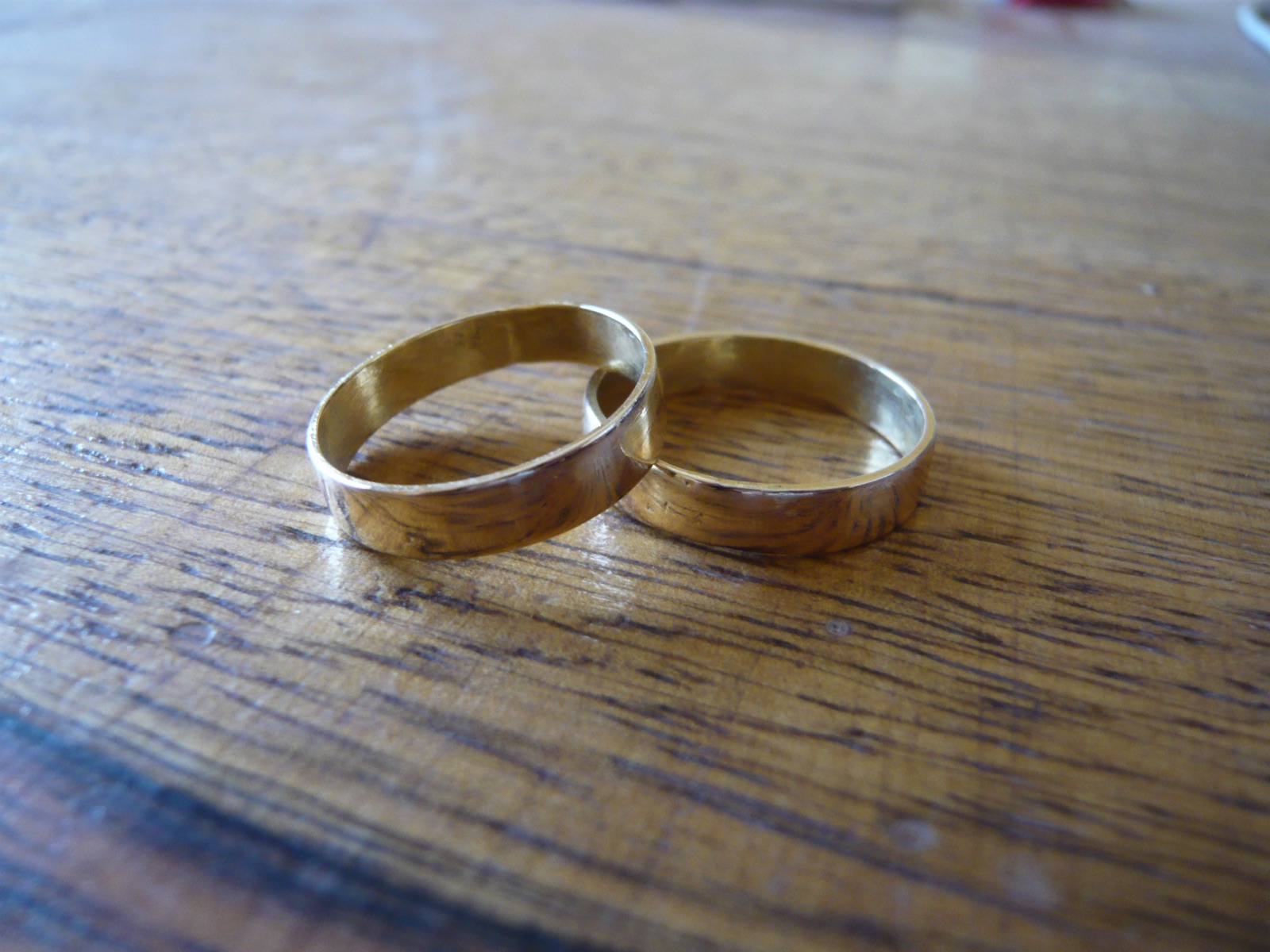 Two worn gold wedding bands resting together on a scuffed oak tabletop — Anna’s thin 1948 band beside the ring it was married alongside in 2019.